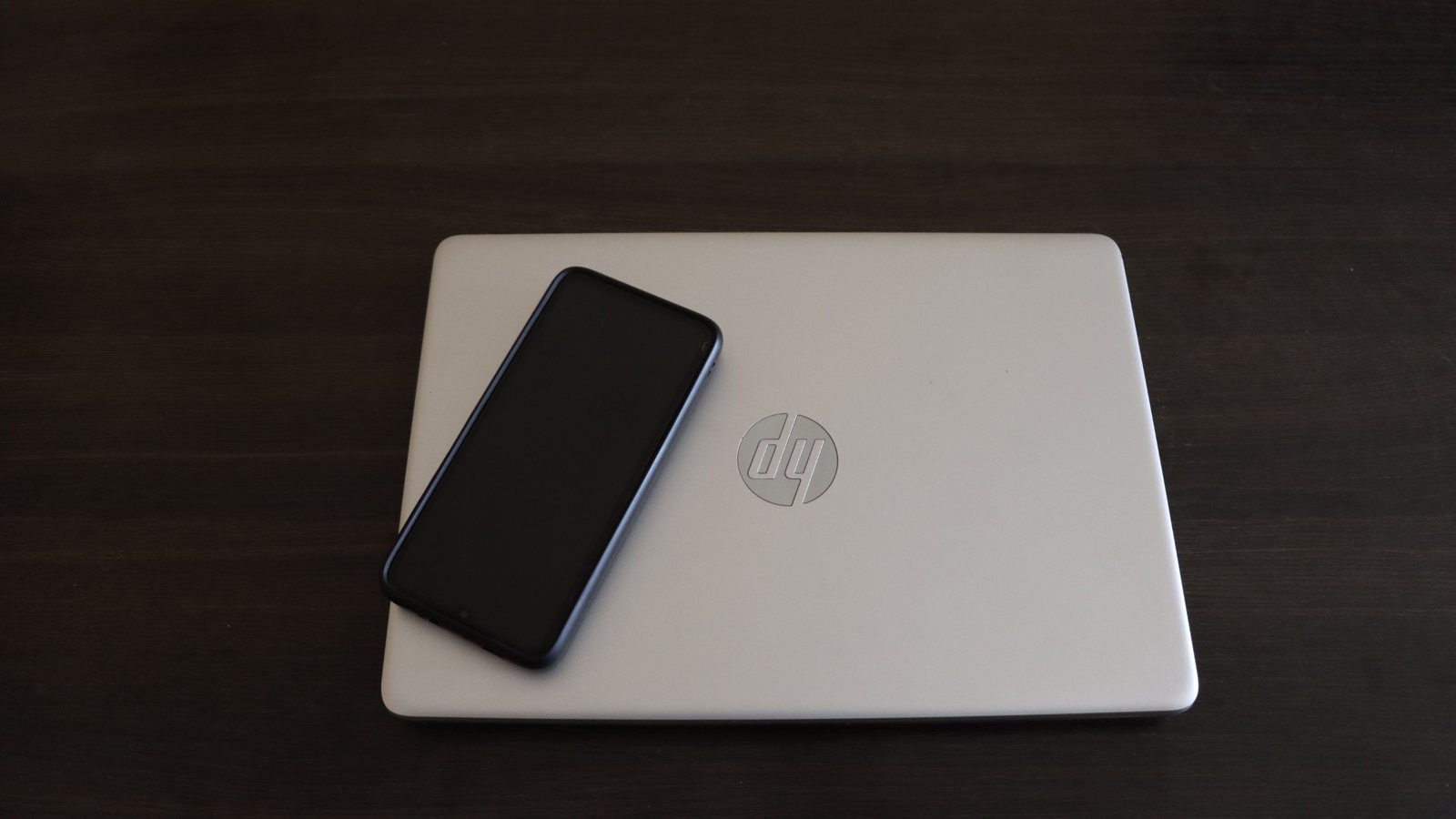 A closed silver laptop with a smartphone resting on top, on a dark wooden desk.