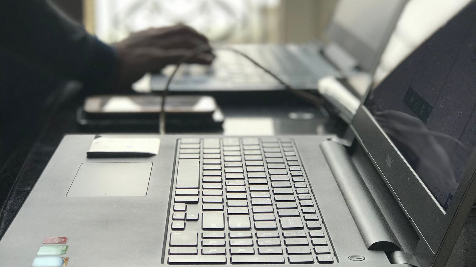 A person typing on a laptop beside a second open laptop, in soft daylight.